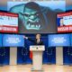 A German politician at a press conference points to a screen labeled “Russian Disinformation,” surrounded by large TV screens broadcasting identical anti-Russia news, while a projected ogre shadow behind him is revealed to come from his own projector.