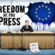 Jurnalis duduk sendirian di depan mikrofon terputus, dengan layar bertuliskan “Freedom of the Press” yang retak dan bendera Uni Eropa yang luntur di belakangnya.